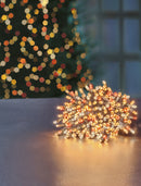 A bundle of warm white LED string lights with red and orange accents coiled on a reflective surface, with a blurred Christmas tree and window in the background.
