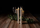 Two tall white taper candles standing lit on a rustic wooden table, surrounded by green garland and small string lights, with wine glasses and more candles blurred in the dark background.