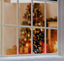 Three black metal star-shaped window decals layered on a white-framed window, with a blurred Christmas tree and warm fireplace in the background.