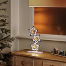 A decorative three-tiered star-shaped table lamp with glowing warm white lights and gold centers, displayed on a wooden cabinet in a cozy room setting next to a Christmas tree.