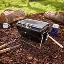 Portable grill on a wooden surface with logs and camping utensils in the background